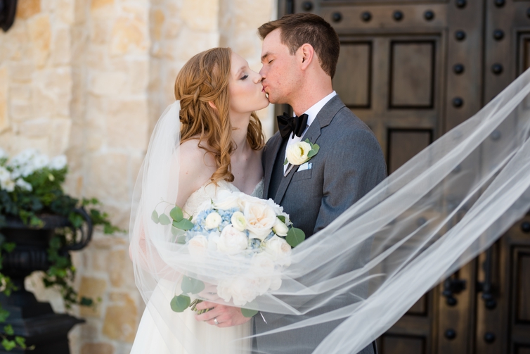 Bride and groom share a kiss while bride holds pink and blue flowers and the veil drapes across the two